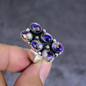 K&ouml;nnte beinhalten: Ein Silberring mit einem Cluster aus ovalen Edelsteinen. Die Steine sind tiefviolett mit goldenen und wei&szlig;en Adern. Der Ring hat ein breites Band. Der Hintergrund ist ein neutrales Grau.