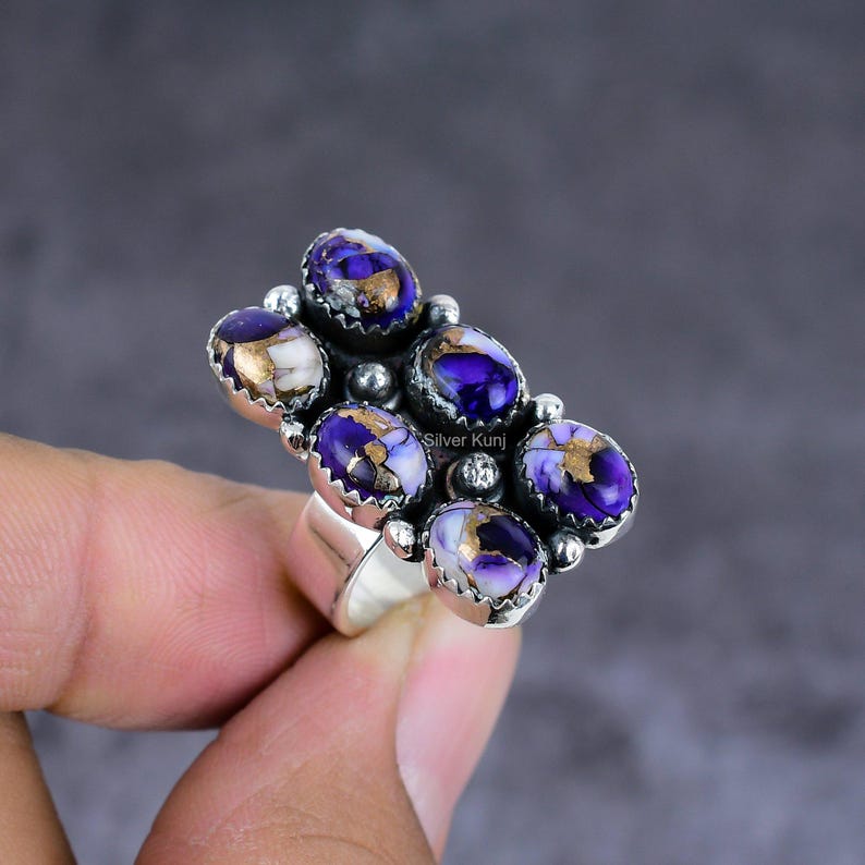 K&ouml;nnte beinhalten: Ein Silberring mit einem Cluster aus ovalen Edelsteinen. Die Steine sind tiefviolett mit goldenen und wei&szlig;en Adern. Der Ring hat ein breites Band. Der Hintergrund ist ein neutrales Grau.