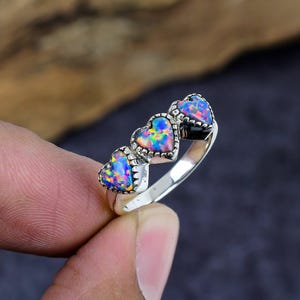 May include: A silver ring featuring three heart-shaped opals. The opals display a vibrant array of colors, including blue, purple, and pink. The ring is held by a person's fingers, showcasing the jewelry's design and details.
