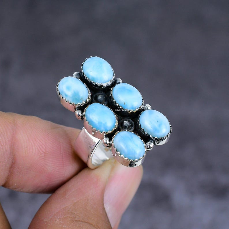 K&ouml;nnte beinhalten: Ein silberner Ring mit sieben ovalen, hellblauen Edelsteinen in einem floralen Muster. Der Ring wird von einer Hand vor einem dunkelgrauen Hintergrund gehalten. Das Ringband ist dick und poliert. Die Steine sind ca. 1 cm lang.