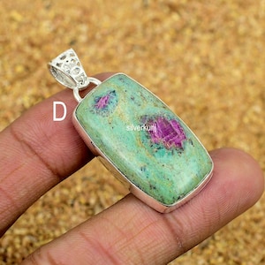 May include: A rectangular pendant with a silver setting. The pendant features a light green stone with vibrant pink inclusions. The top of the pendant has a decorative silver bail. The word "D" is visible on the left side of the pendant.