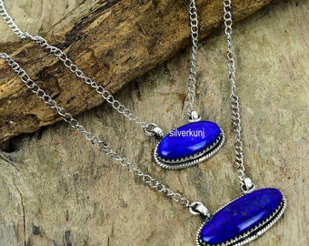 May include: Two silver chains with oval lapis lazuli pendants. The pendants have a silver border with a decorative design. The chains are layered on a wooden surface.