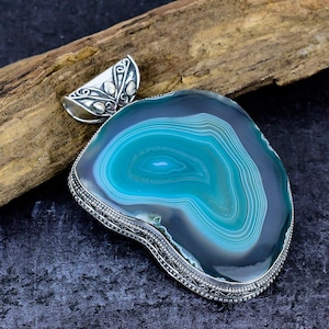 May include: A pendant featuring a large, polished, teal and dark gray agate slice with concentric banding. The stone is encased in a decorative silver setting with intricate metalwork along the edges and a matching ornate silver bail at the top.