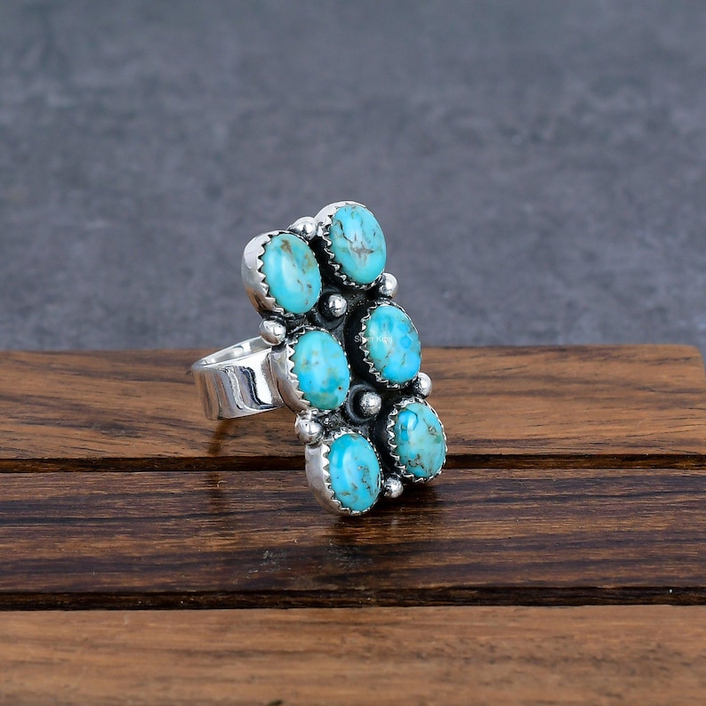 K&ouml;nnte beinhalten: Ein silberner Ring mit sechs ovalen t&uuml;rkisfarbenen Steinen in vertikaler Anordnung. Jeder Stein hat einen detaillierten Silberrahmen. Der Ring wird auf einer Holzoberfl&auml;che pr&auml;sentiert. Das Design des Rings erinnert an eine Traube.