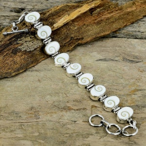 Puede incluir: Un brazalete de plata con un diseño de cadena de eslabones y abalorios ovalados de concha blanca con patrones de espiral marrón. El brazalete tiene un cierre de langosta.