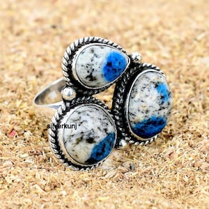 Op de afbeelding: Zilveren ring met drie ovale en druppelvormige stenen. De stenen hebben een wit en grijs gespikkeld patroon met blauwe accenten. De ring ligt op een bedje van bruine houtkrullen.