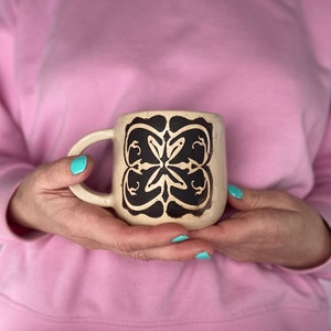 Peut inclure: Une tasse en céramique beige avec un motif géométrique noir. La tasse a une anse et est tenue par les mains d'une personne.