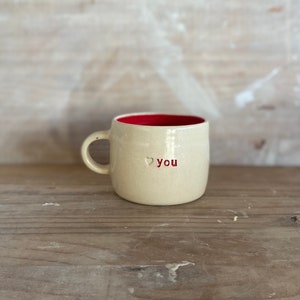 Puede incluir: Taza de cerámica color crema con interior rojo. La taza tiene un asa pequeña y las palabras "❤ you" en rojo. La taza está sobre una superficie de madera.