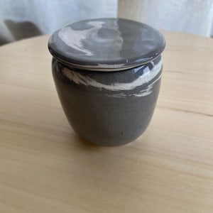 May include: A small, grey ceramic jar with a lid. The jar has a white marbled design and a glossy finish.