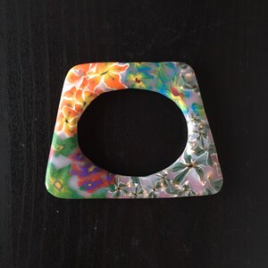 May include: A white, square-shaped bangle with a floral design. The flowers are painted in shades of orange, yellow, green, blue, and purple.