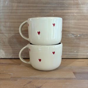 May include: Two white ceramic mugs with red heart designs. The mugs are stacked on top of each other.