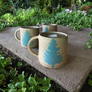 May include: Three ceramic mugs with blue tree designs sit on a stone patio. The mugs are filled with a light-colored beverage and ice.