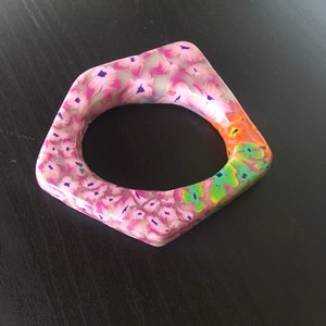 May include: A square-shaped bangle bracelet with a floral pattern. The bracelet features pink flowers with purple accents on a light background. One section of the bracelet has a colorful design with orange, green, and yellow hues.