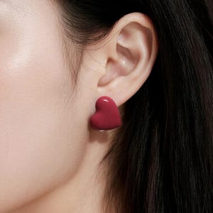 Könnte beinhalten: Nahaufnahme eines Ohrs mit einem roten herzförmigen Ohrstecker. Der Ohrring hat eine glänzende, rote Farbe und befindet sich am Ohrläppchen. Dunkles Haar ist im Hintergrund zu sehen.