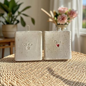 Può includere: Due targhe rettangolari in ceramica color bianco sporco con texture maculata e ganci metallici per appenderle. Una targa riporta il nome Greta e un'impronta di zampa, l'altra il nome Tato e un piccolo simbolo di un cuore rosso.