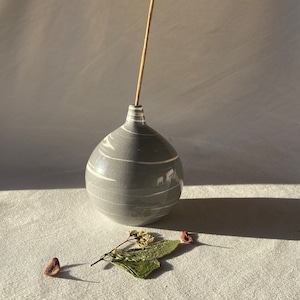 Può includere: Un porta incenso in ceramica grigia con striature bianche e una glassa craquelata. Un bastoncino di incenso in legno è posto nel porta incenso.