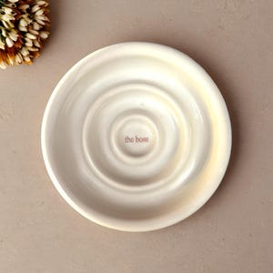 Può includere: Piatto in ceramica bianco sporco con cerchi concentrici e la scritta "the boss" al centro. Il piatto è rotondo e ha un bordo leggermente rialzato. Un piccolo bouquet di fiori secchi è nell'angolo in alto a sinistra.