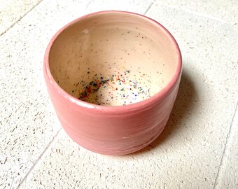 May include: A handmade ceramic bowl with a soft pink exterior and a speckled interior. The bowl has a rounded shape and a smooth, matte finish. The interior is speckled with colorful flecks.