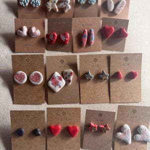 Può includere: Una collezione di orecchini a bottone fatti a mano esposti su cartoncini marroni. Gli orecchini presentano vari design, tra cui cuori, stelle, fiori e forme astratte, in colori come rosso, rosa, blu e bianco. Alcuni cartoncini riportano il testo "AIR" e "Barcelona".
