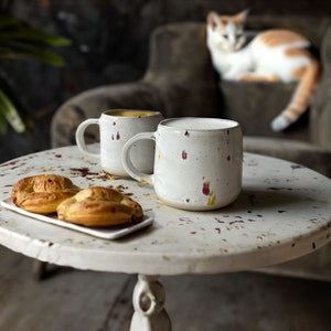 Può includere: Due tazze in ceramica bianca con dettagli di vernice maculata rossa e gialla sono su un tavolo rotondo bianco usurato. Due pasticcini cotti su un piatto rettangolare bianco. Un gatto riposa su una poltrona marrone sullo sfondo.