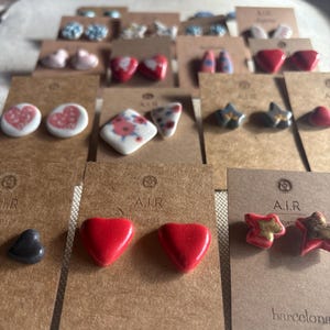 Könnte beinhalten: Eine Sammlung handgefertigter Ohrstecker, präsentiert auf braunen Karten. Die Ohrringe weisen verschiedene Designs auf, darunter Herzen, Sterne und Blumenmuster, in Farben wie Rot, Weiß und Blau. Auf den Karten steht der Text "A.I.R" und "barcelona".