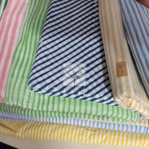 Może przedstawiać: Stos materiału w paski w różnych kolorach, w tym różowym, zielonym, niebieskim i żółtym. Materiał jest starannie złożony i ma białe tło.