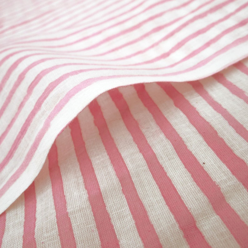 May include: Pink and white striped fabric with a soft, linen texture. The fabric is folded over, creating a wave-like pattern.