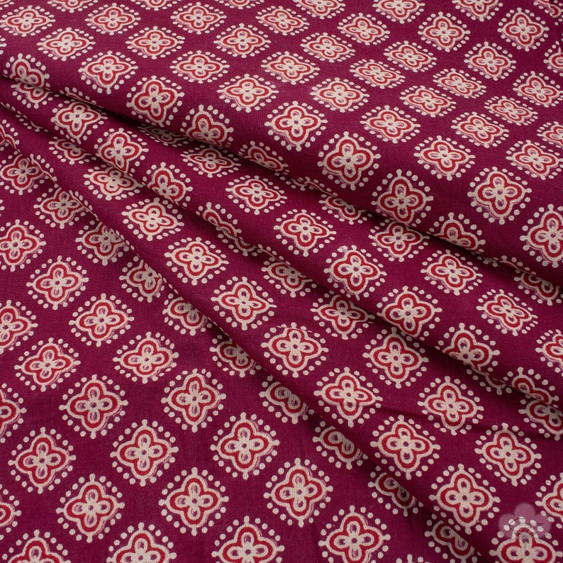 May include: A close-up of a folded fabric with a repeating pattern. The fabric is a deep burgundy color, with a pattern of white and red floral-like shapes. The pattern is evenly spaced across the fabric.
