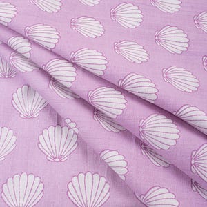 Pode incluir: Um tecido lilás claro com um padrão repetido de ilustrações de conchas brancas. O tecido está dobrado, mostrando a textura e o design. As conchas são detalhadas com linhas curvas.