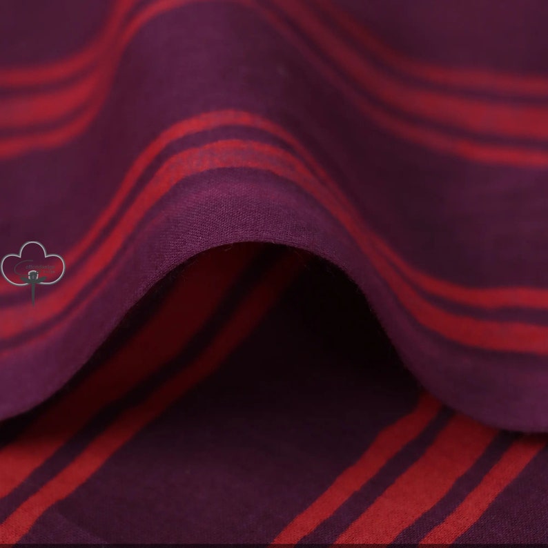 May include: Close-up of a fabric with a deep purple base and bold red stripes. The stripes are uneven and wavy, creating a dynamic pattern. The fabric appears soft and flowing, with visible folds and shadows.