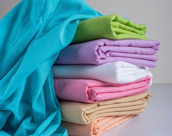 Puede incluir: Una pila de tela doblada en varios colores, incluyendo turquesa, verde, morado, blanco, rosa y beige. La tela parece suave y ligera, posiblemente lino o algodón, y está cuidadosamente doblada.