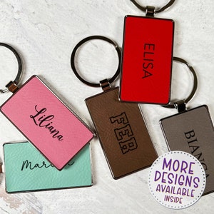 May include: Rectangular keychains in various colors, including red, pink, and brown, with personalized names and initials. The keychains have silver-colored metal rings. The image also includes a circular graphic that says "MORE DESIGNS AVAILABLE INSIDE."