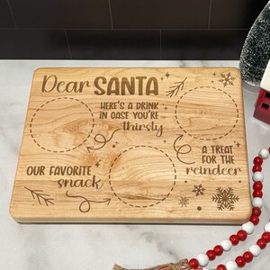 May include: A wooden cutting board with the text "Dear Santa" and three circles for placing treats. The circles are labeled "Our Favorite Snack", "Here's a Drink in Case You're Thirsty", and "A Treat for the Reindeer".