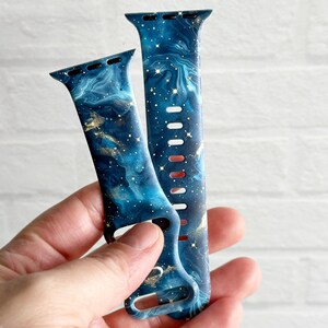 May include: Two blue and gold galaxy-print Apple Watch bands. The bands have a marble-like design with gold flecks and a dark blue background.