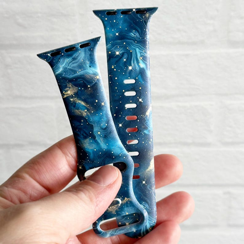 May include: Two blue and gold galaxy-print Apple Watch bands. The bands have a marble-like design with gold flecks and a dark blue background.