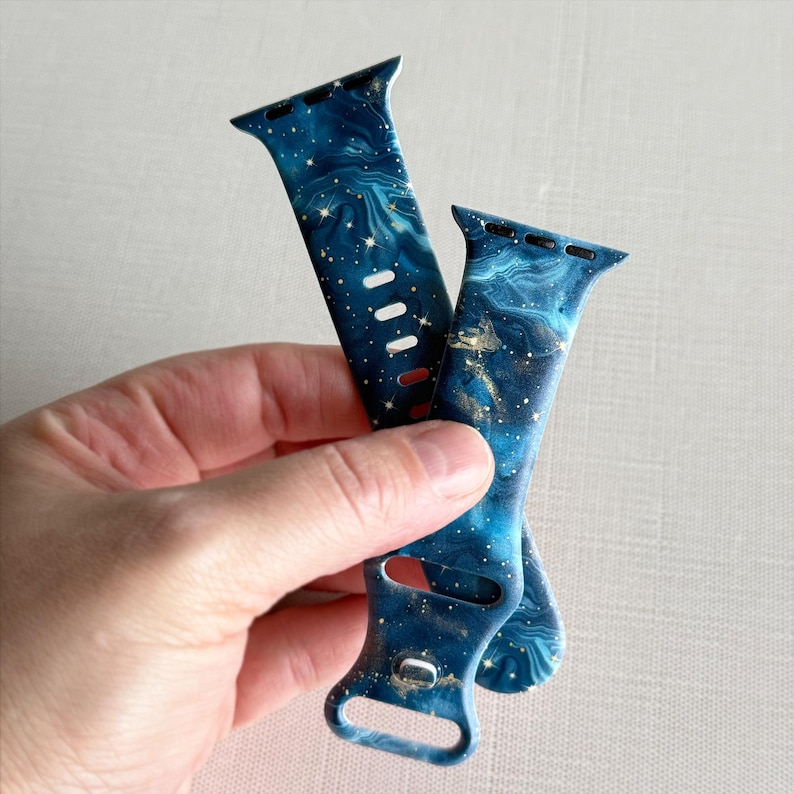 May include: Two blue and gold galaxy print Apple Watch bands. The bands are made of silicone and have a marbled effect with gold glitter.