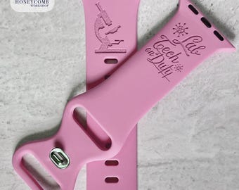 May include: Pink Apple Watch bands with engraved designs. One band features a microscope, the other has the text "Lab Tech on Duty" with a graphic. The bands are made of a flexible material and have a silver clasp.