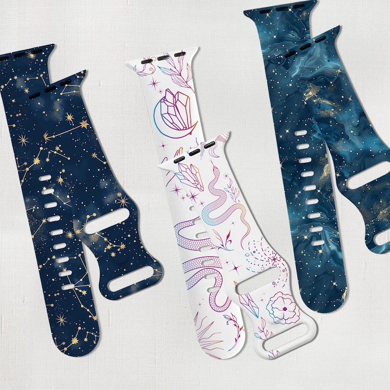 May include: Three different Apple Watch bands with celestial and nature-inspired designs. The first band features a dark blue background with gold constellations and stars. The second band has a white background with a colorful snake, crystals, and flowers. The third band has a dark blue and gold marble pattern.