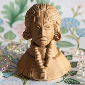 Puede incluir: Un busto de color marrón claro de una figura con rasgos faciales detallados, incluyendo ojos cerrados y un tocado estampado. La figura tiene un collar y una base redondeada. El fondo presenta un estampado floral en colores pastel.