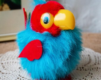 May include: A plush toy parrot with a red head, blue body, and yellow beak. The bird has large, cartoonish eyes and red feet. The toy is made of soft, fuzzy material and is standing on a white, lacy surface.