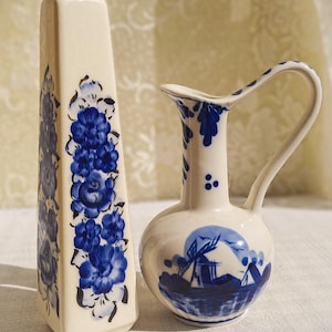 Könnte beinhalten: Zwei cremefarbene Keramikvasen mit blauen Blumen- und Landschaftsdesigns. Die größere Vase ist rechteckig mit blauen Blumenmustern. Die kleinere Vase hat einen Henkel und eine Windmühlen-Szene.