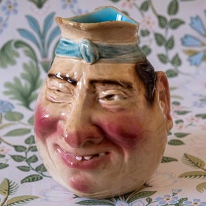 May include: A novelty ceramic jug in the shape of a smiling face, with rosy cheeks and a winking eye. The jug is tan with brown hair and a blue band around the top. The interior of the jug is light blue.
