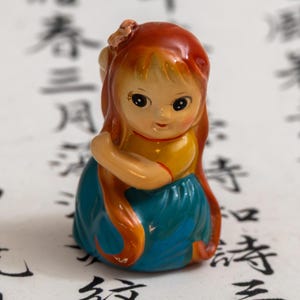 Peut inclure: Petite figurine vintage représentant une jeune fille aux longs cheveux roux. Elle porte un haut jaune et une jupe bleue. La figurine a un visage peint et doux, et tient ses bras près de son corps. Le fond présente de la calligraphie asiatique.