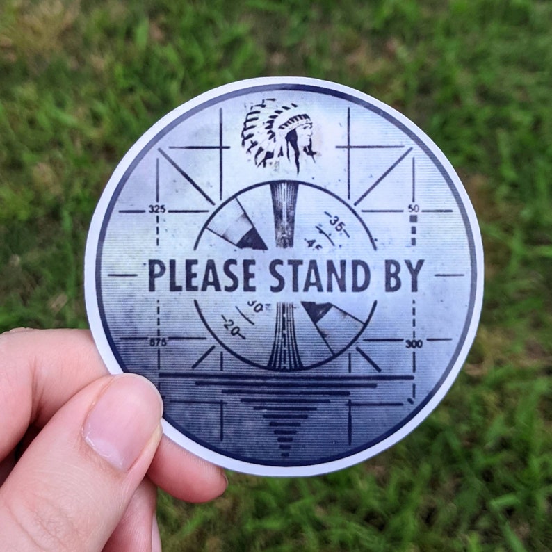May include: A round sticker with a vintage television test pattern design. The sticker features the text "PLEASE STAND BY" in bold, white letters. An illustration of a Native American headdress is at the top. The background is a gradient of gray and blue.