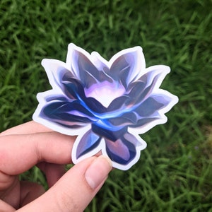 May include: A sticker featuring a stylized lotus flower design. The flower has a gradient of colors, including purple, blue, and white, with a glowing effect in the center. The sticker is held against a backdrop of green grass.