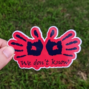 May include: A sticker with a red handprint design and the text "We don't know" in a handwritten style. The handprints are black and the sticker has a white border. The sticker is on a green grassy background.