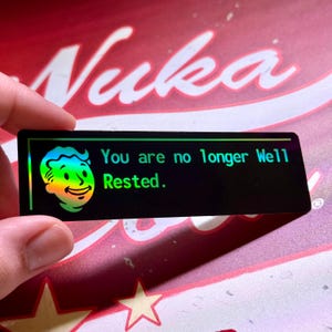 May include: A black rectangular sticker with the text "You are no longer Well Rested." and a cartoon character with a rainbow-colored head. The sticker has a holographic effect, with the background featuring the word "Nuka".