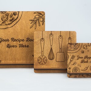 May include: Three wooden recipe book stands of different sizes. The largest stand has the text "Your Recipe Book Lives Here" and a pizza graphic. The middle stand has a graphic of kitchen utensils. The smallest stand has a graphic of various food items and the text "Your Recipe Book".