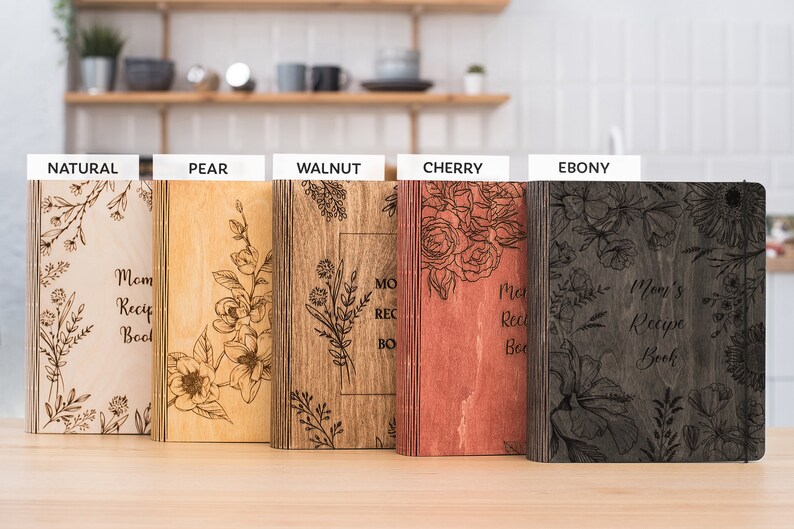 Puede incluir: Cinco libros de recetas de madera con dise&ntilde;os florales y el texto "Mom's Recipe Book" en diferentes manchas de madera: natural, pera, nogal, cerezo y &eacute;bano.