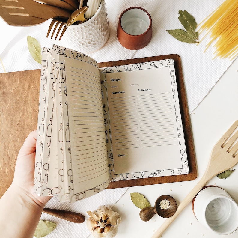 Peut inclure: Un livre de recettes ouvert avec des pages vierges pour &eacute;crire des recettes. Le livre a une couverture brune et est d&eacute;cor&eacute; d'une illustration en noir et blanc d'articles de cuisine.